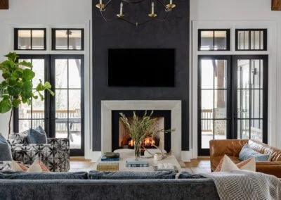 A beautifully designed living room featuring a cozy fireplace with a stone surround, flanked by large black-framed windows. The room is furnished with a blue velvet sofa, a leather chair, and patterned cushions. A glass coffee table with decorative books and a vase adds to the stylish decor. This design exemplifies the high-quality craftsmanship provided by Sixteen Penny Construction in custom home building.