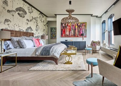A sophisticated bedroom featuring a wooden bed with luxurious grey bedding and vibrant pillows, complemented by a striking chandelier. Colorful artwork adorns the walls, and chic furniture, including armchairs and a decorative rug, creates a welcoming atmosphere. Large windows flood the room with natural light. This room exemplifies the work of luxury home builders in Charlotte, showcasing high-end design and exceptional craftsmanship.