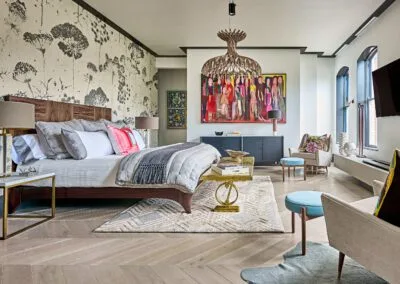 A sophisticated bedroom featuring a wooden bed with luxurious grey bedding and vibrant pillows, complemented by a striking chandelier. Colorful artwork adorns the walls, and chic furniture, including armchairs and a decorative rug, creates a welcoming atmosphere. Large windows flood the room with natural light. This room exemplifies the work of luxury home builders in Charlotte, showcasing high-end design and exceptional craftsmanship.
