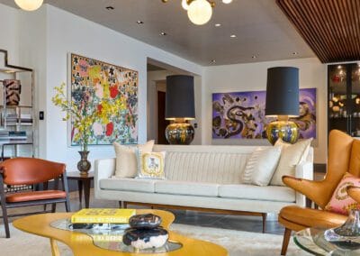A stylish living room featuring a sleek sofa, a vibrant chandelier, and captivating artwork. The space is elegantly designed with mid-century modern furniture, including a distinctive coffee table and armchairs. The room offers a perfect blend of comfort and contemporary aesthetics. Get in Touch for more information on creating your dream home.